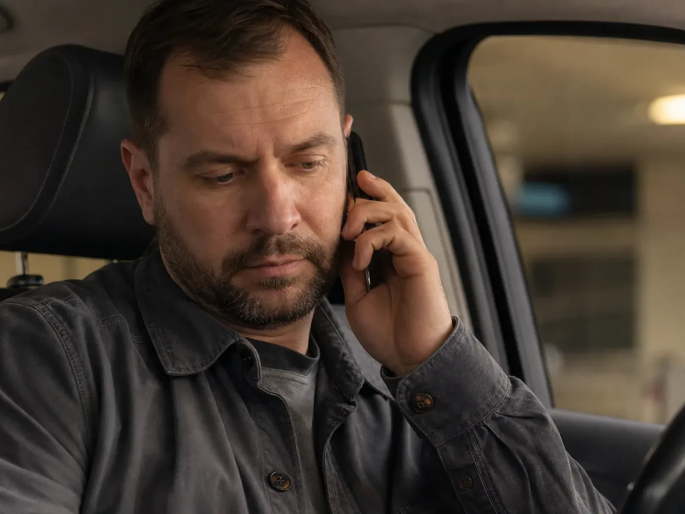 A person sits in a parked car holding a mobile phone to their ear, with a steering wheel visible and a blurred indoor parking area in the background