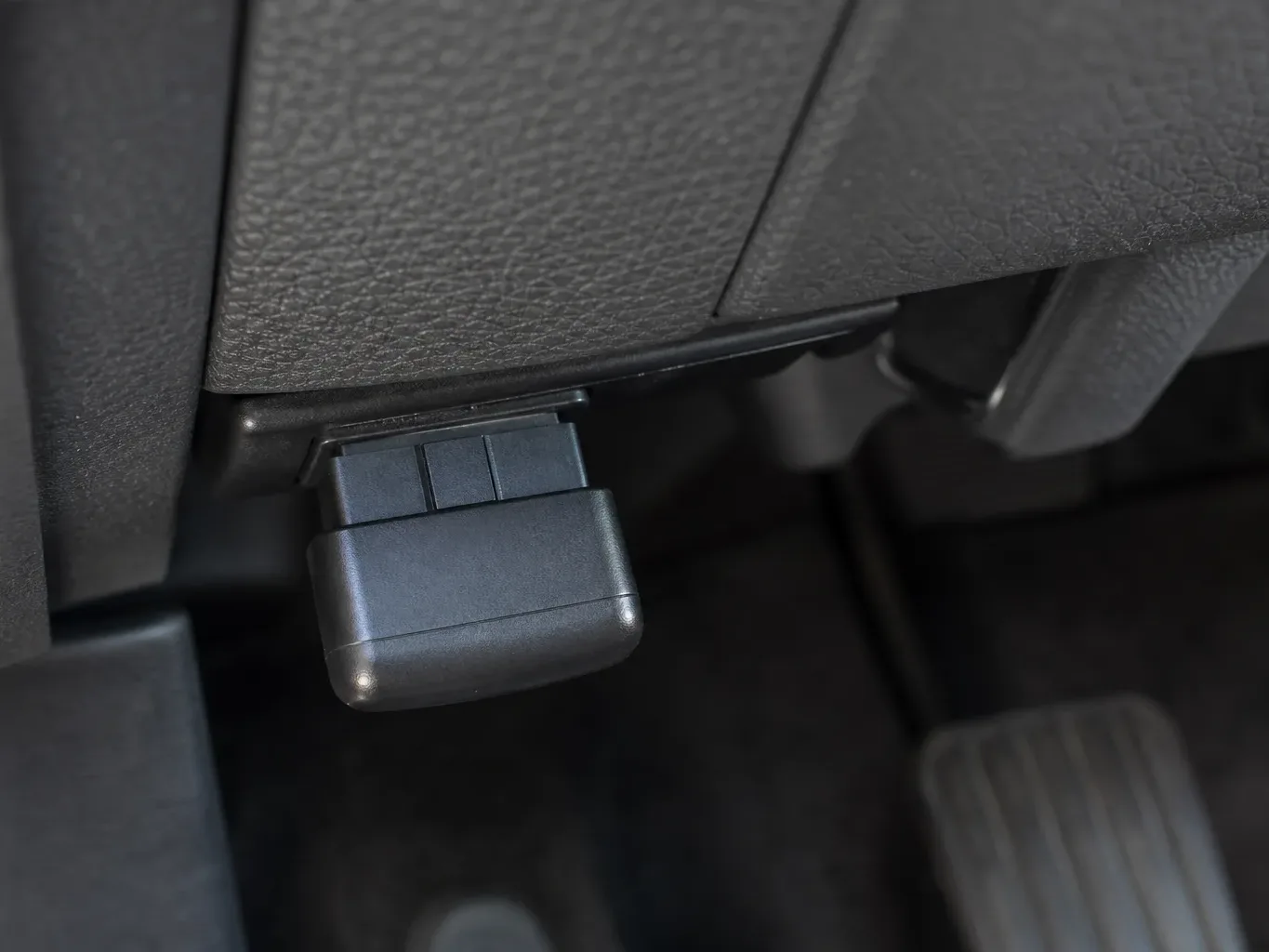 A small black device plugged into a port beneath a car dashboard, with pedals visible in the background
