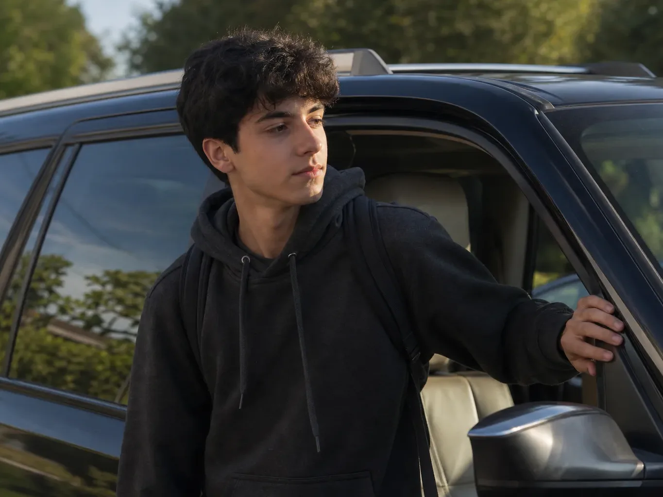 A person in a dark hoodie stands at the open door of a dark SUV, one hand resting on the door frame, looking to one side