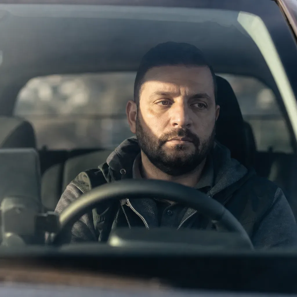 A person sits behind the steering wheel of a vehicle, looking ahead through the windscreen in cool daylight