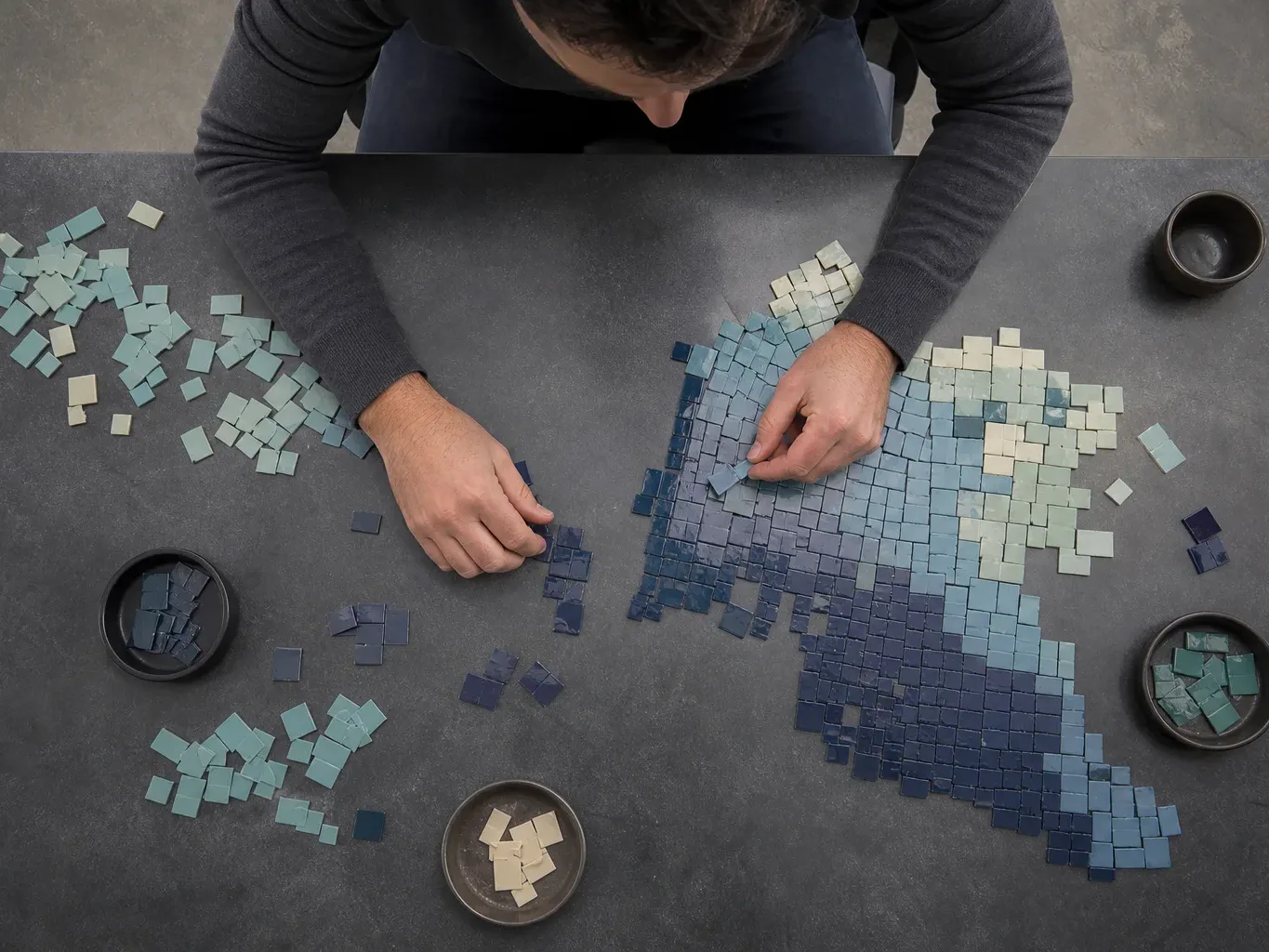 A person arranges small blue and white square tiles on a dark surface to form a mosaic pattern, viewed from directly overhead