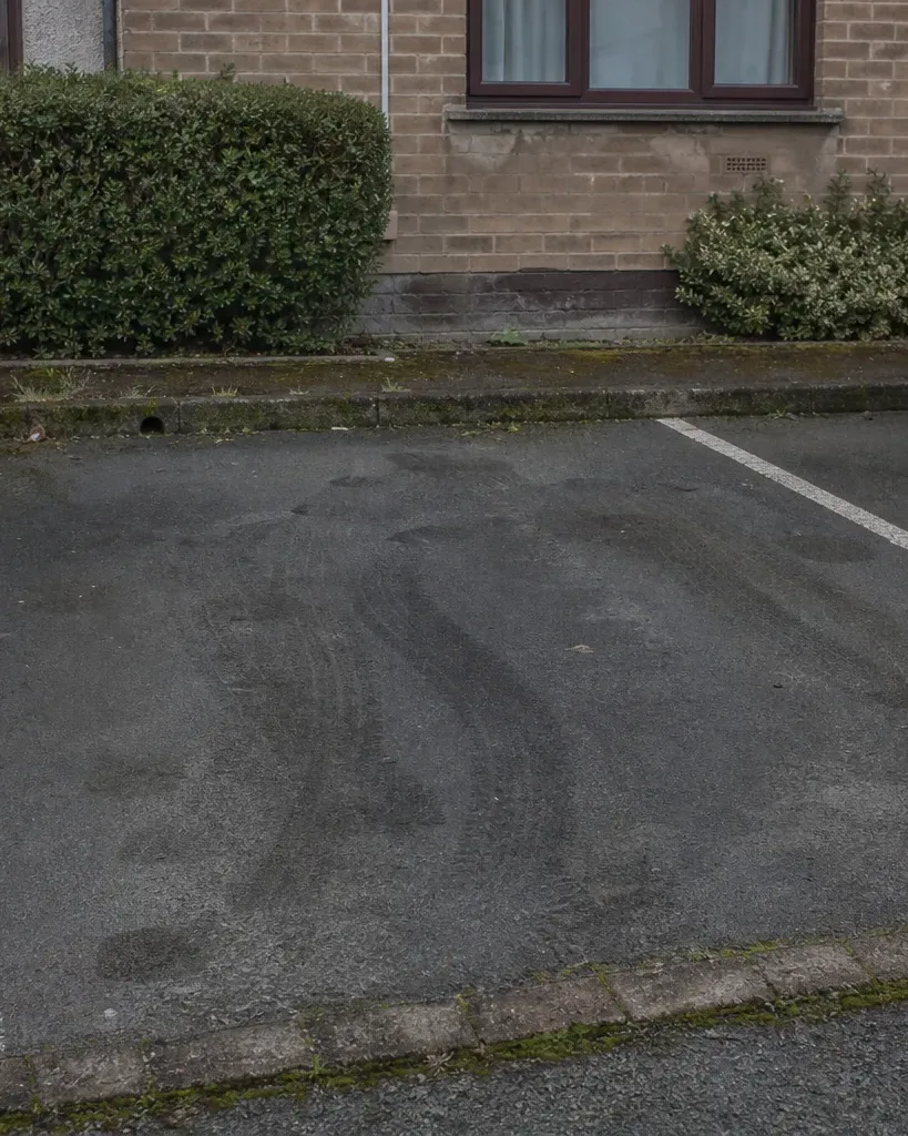 An empty parking bay between two parked cars outside a residential brick building, with oil stains and tyre marks visible on the wet asphalt
