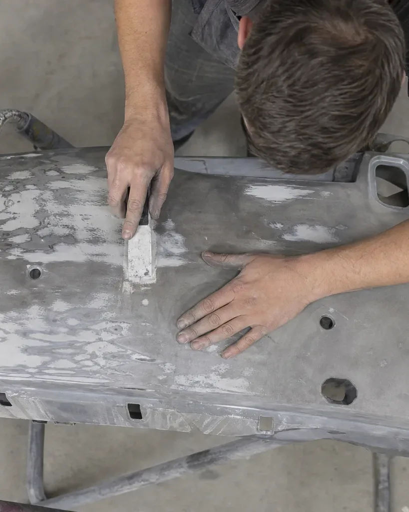 A person bends over a stripped car body panel on a workshop stand, smoothing filler with a hand tool, viewed from directly overhead