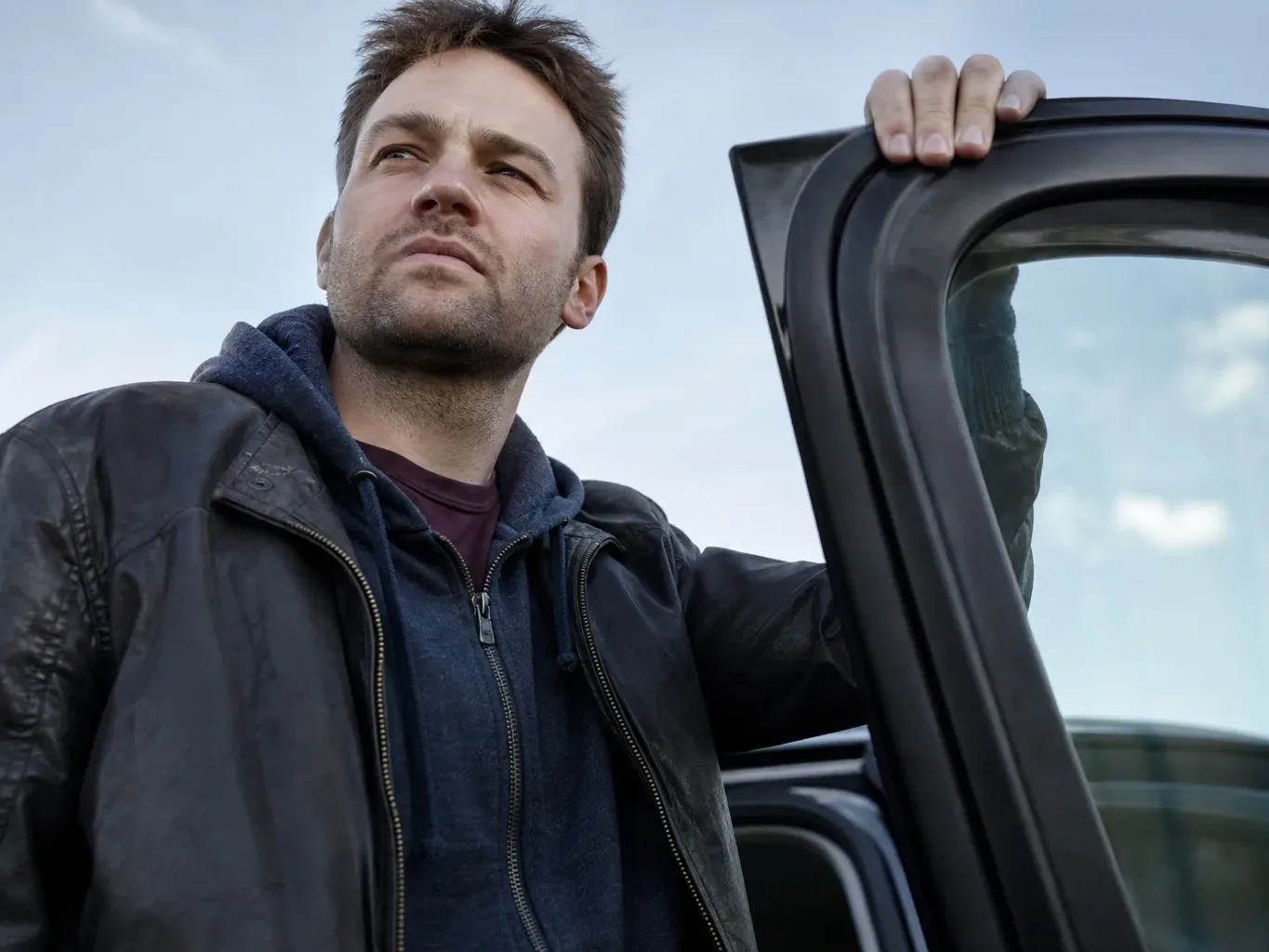 A person in a leather jacket holds the open door frame of a vehicle, looking away into the distance against a pale cloudy sky