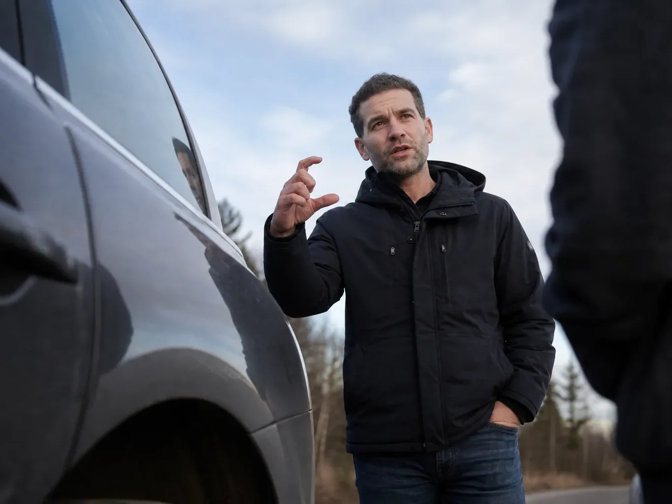 A person in a dark jacket stands beside a parked vehicle in cool daylight, one hand raised with fingers gesturing as they speak to someone out of frame