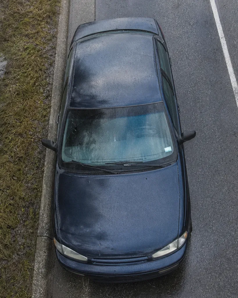 A dark blue saloon car is parked on a kerb beside wet grass, photographed from directly above on an overcast day
