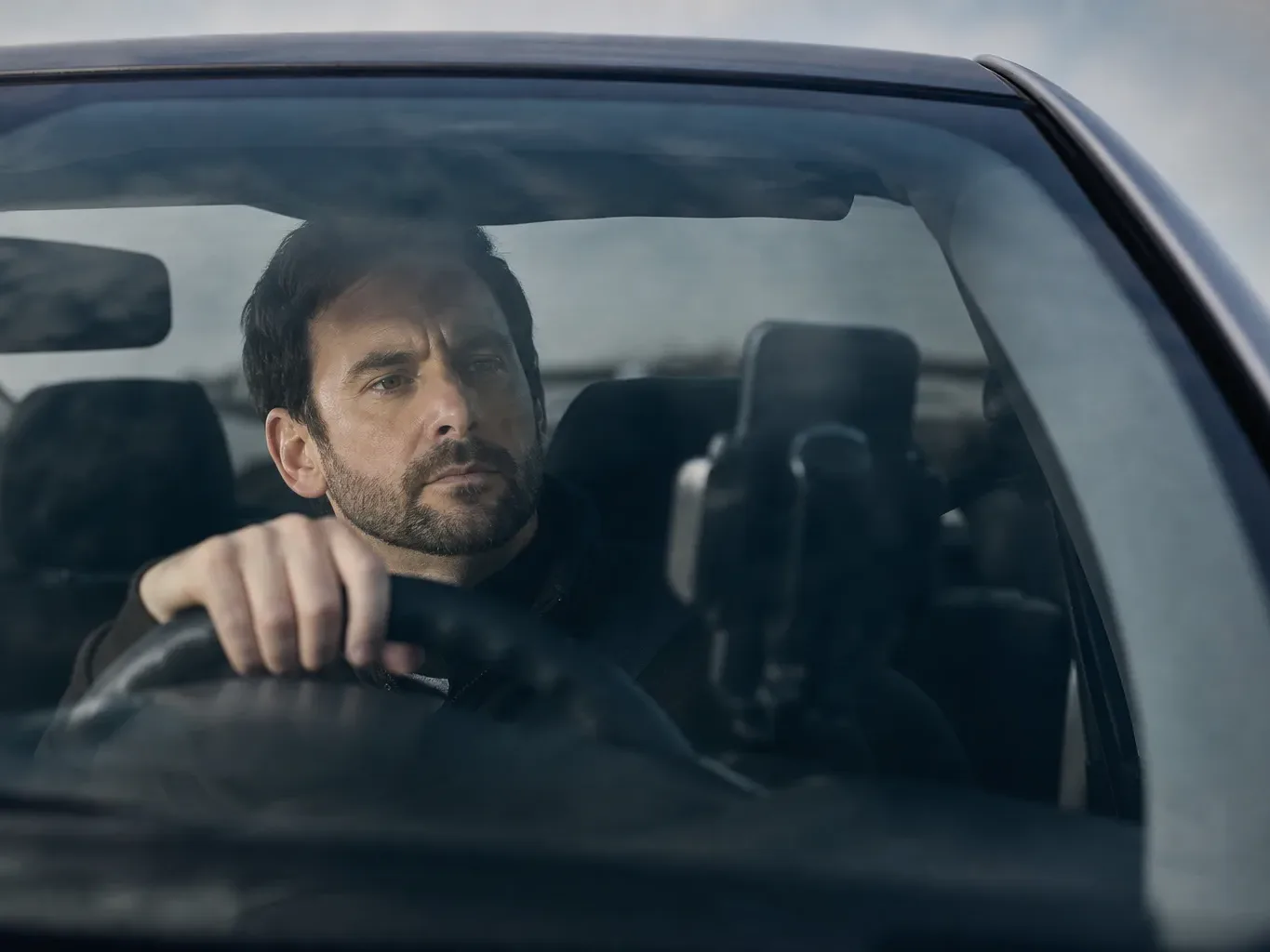 A person sits in the driver's seat of a car, one hand resting on the steering wheel, gazing ahead through the windscreen in overcast daylight