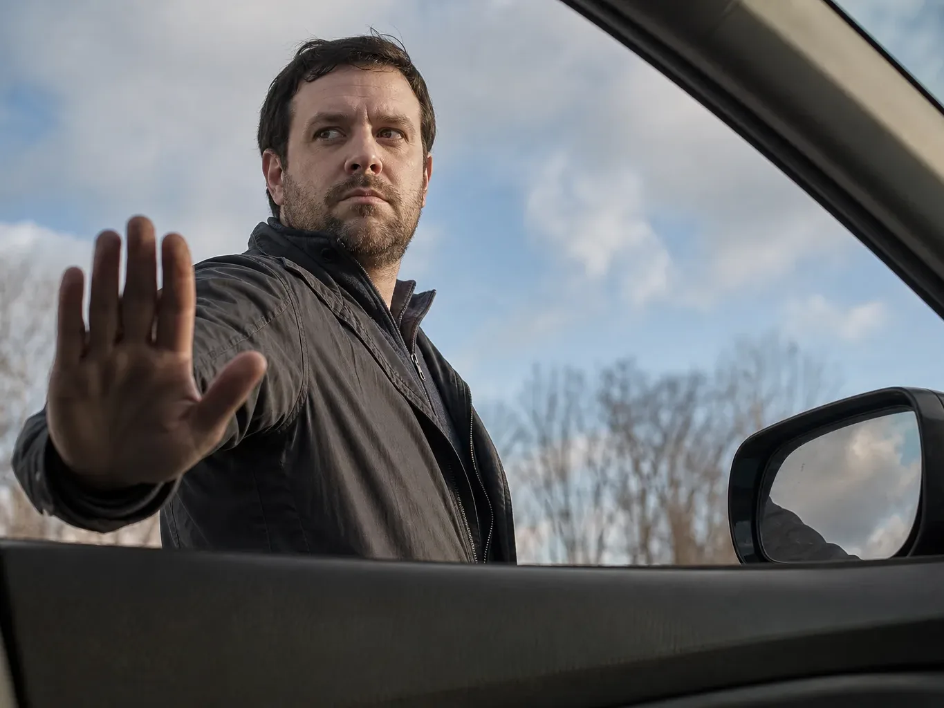 A person stands outside a car door, one hand held out flat in a stopping gesture, looking sideways against a cloudy sky